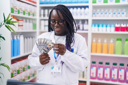 Foto de Farmacéutica afroamericana sonriendo confiada contando dólares en farmacia - Imagen libre de derechos
