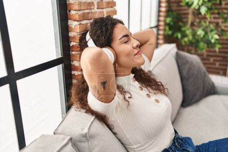 Téléchargez les photos : Jeune belle femme hispanique écoutant de la musique assise sur le canapé à la maison - en image libre de droit