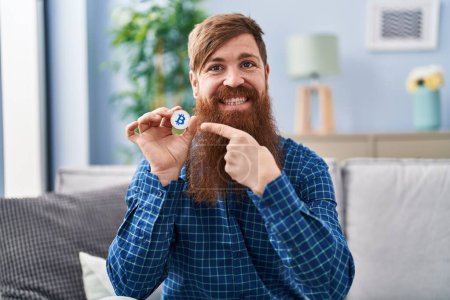 Foto de Hombre caucásico con barba larga sosteniendo moneda virtual bitcoin sonriendo feliz señalando con la mano y el dedo - Imagen libre de derechos