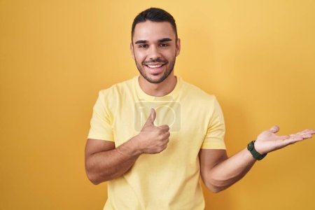 Téléchargez les photos : Jeune homme hispanique debout sur fond jaune montrant la main de la paume et faisant un geste correct avec les pouces levés, souriant heureux et joyeux - en image libre de droit