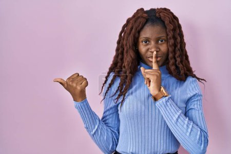 Foto de Mujer africana de pie sobre fondo rosado pidiendo estar callada con el dedo en los labios apuntando con la mano hacia un lado. silencio y concepto secreto. - Imagen libre de derechos
