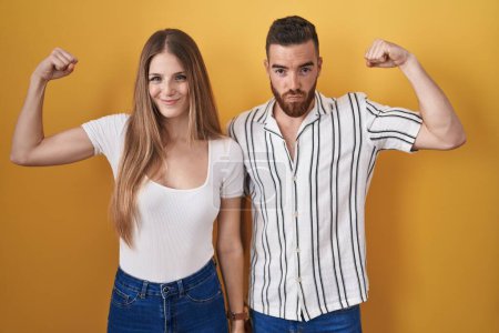 Foto de Young couple standing over yellow background strong person showing arm muscle, confident and proud of power - Imagen libre de derechos