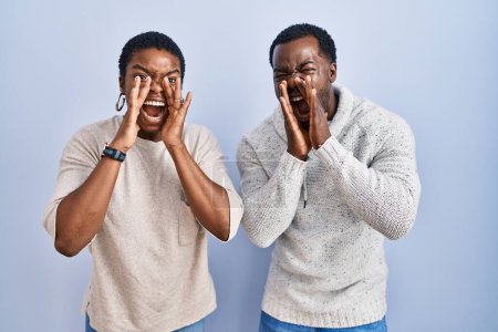 Téléchargez les photos : Jeune couple afro-américain debout sur fond bleu ensemble criant en colère à haute voix avec les mains sur la bouche - en image libre de droit