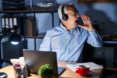 Photo for Hispanic senior man wearing call center agent headset at night shouting and screaming loud to side with hand on mouth. communication concept. - Royalty Free Image