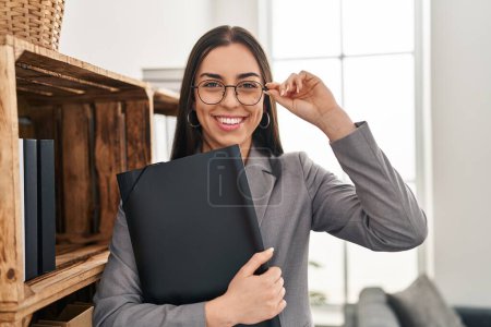 Photo for Young beautiful hispanic woman psychologist smiling confident holding binder at psychology clinic - Royalty Free Image