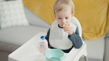 Photo for Adorable blond toddler sitting on highchair eating snack at home - Royalty Free Image
