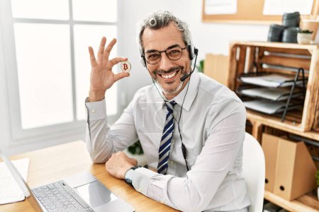 Foto de Hombre de pelo gris de mediana edad agente del centro de llamadas sonriendo confiado sosteniendo bitcoin en la oficina - Imagen libre de derechos