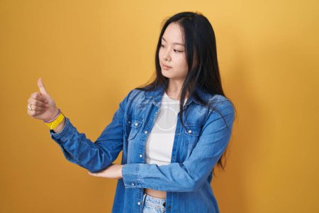 Téléchargez les photos : Young asian woman standing over yellow background looking proud, smiling doing thumbs up gesture to the side - en image libre de droit