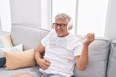Foto de Middle age grey-haired man listening to music doing guitar gesture at home - Imagen libre de derechos