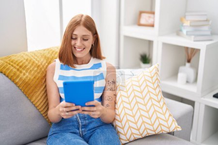 Téléchargez les photos : Jeune rousse femme en utilisant touchpad assis sur le canapé à la maison - en image libre de droit