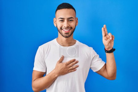 Téléchargez les photos : Jeune homme hispanique debout sur fond bleu souriant jurer avec la main sur la poitrine et les doigts vers le haut, faisant un serment de fidélité promesse - en image libre de droit