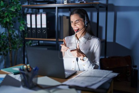 Photo for Young brunette woman wearing call center agent headset working late at night pointing fingers to camera with happy and funny face. good energy and vibes. - Royalty Free Image
