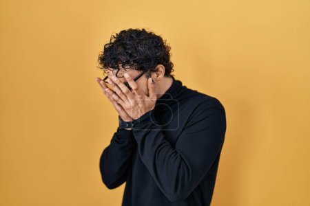 Foto de Hispanic man standing over yellow background with sad expression covering face with hands while crying. depression concept. - Imagen libre de derechos
