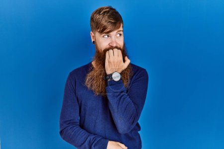 Foto de Redhead man with long beard wearing casual blue sweater over blue background looking stressed and nervous with hands on mouth biting nails. anxiety problem. - Imagen libre de derechos