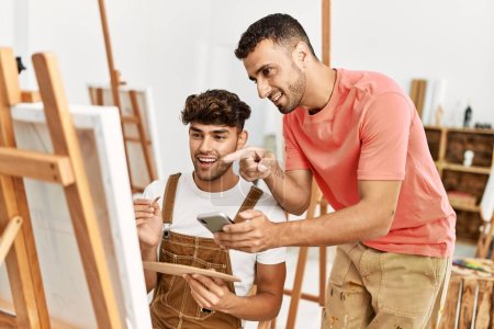 Foto de Dos hombres hispanos en pareja sonriendo confiados usando un smartphone y dibujando en un estudio de arte - Imagen libre de derechos