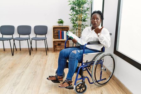 Photo for Young black woman sitting on wheelchair at waiting room surprised pointing with hand finger to the side, open mouth amazed expression. - Royalty Free Image