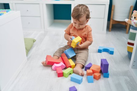 Foto de Adorable niño rubio jugando con bloques de geometría sentado en el suelo en el jardín de infantes - Imagen libre de derechos