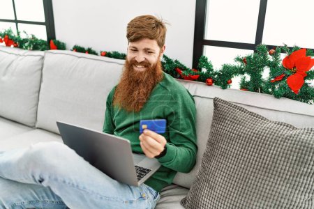 Foto de Young redhead man using laptop and credit card sitting by christmas decor at home - Imagen libre de derechos