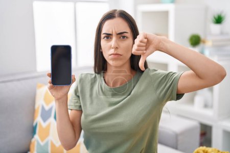 Photo for Young brunette woman holding smartphone showing blank screen with angry face, negative sign showing dislike with thumbs down, rejection concept - Royalty Free Image