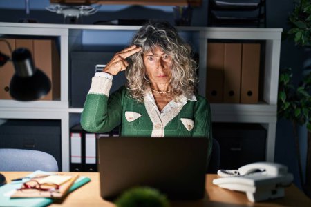 Foto de Mujer de mediana edad que trabaja de noche usando computadora portátil disparando y matándose apuntando de la mano y los dedos a la cabeza como arma, gesto suicida. - Imagen libre de derechos