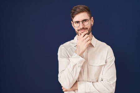 Photo for Hispanic man with beard standing over blue background looking confident at the camera with smile with crossed arms and hand raised on chin. thinking positive. - Royalty Free Image
