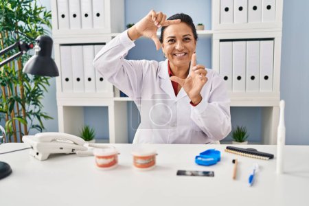 Photo for Hispanic mature dentist woman working at the clinic smiling making frame with hands and fingers with happy face. creativity and photography concept. - Royalty Free Image