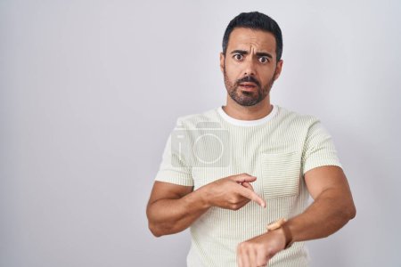 Téléchargez les photos : Hispanic man with beard standing over isolated background in hurry pointing to watch time, impatience, upset and angry for deadline delay - en image libre de droit