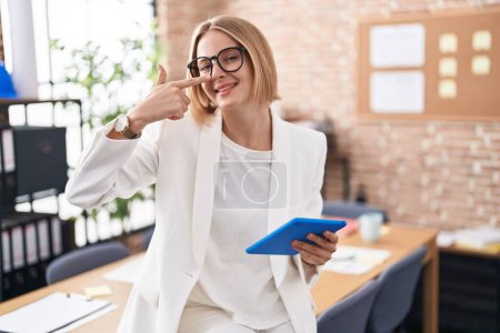 Foto de Young caucasian woman working at the office wearing glasses pointing with hand finger to face and nose, smiling cheerful. beauty concept - Imagen libre de derechos
