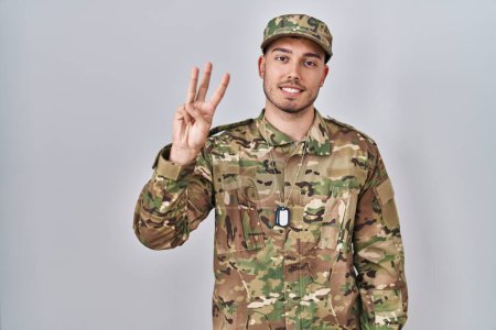 Foto de Joven hombre hispano usando uniforme camuflaje del ejército mostrando y señalando hacia arriba con los dedos número tres mientras sonríe confiado y feliz. - Imagen libre de derechos