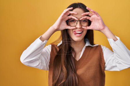 Photo for Young brunette woman standing over yellow background wearing glasses doing ok gesture like binoculars sticking tongue out, eyes looking through fingers. crazy expression. - Royalty Free Image