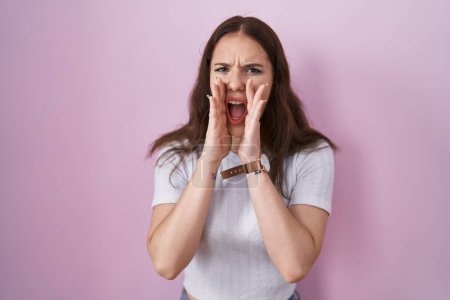 Foto de Joven chica hispana de pie sobre fondo rosa gritando enojado en voz alta con las manos sobre la boca - Imagen libre de derechos