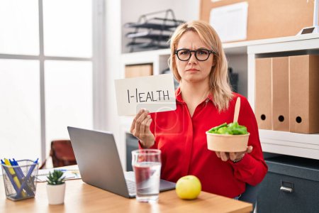 Foto de Mujer rubia que trabaja en la oficina comiendo comida saludable expresión despistada y confusa. concepto de duda. - Imagen libre de derechos