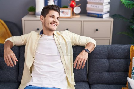 Téléchargez les photos : Jeune homme hispanique souriant confiant assis sur le canapé à la maison - en image libre de droit