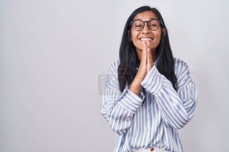 Foto de Joven mujer hispana usando gafas rezando con las manos juntas pidiendo perdón sonriendo confiada. - Imagen libre de derechos