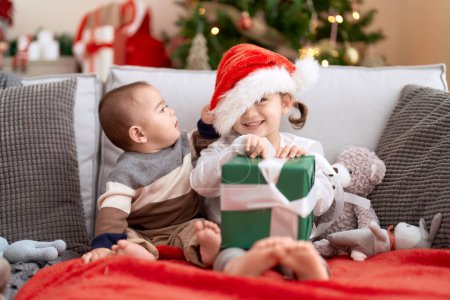 Photo for Brother and sister opening gift sitting on sofa by christmas tree at home - Royalty Free Image