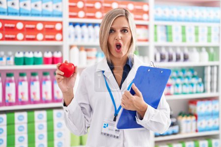 Téléchargez les photos : Young woman working at pharmacy drugstore holding heart afraid and shocked with surprise and amazed expression, fear and excited face. - en image libre de droit