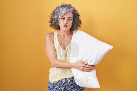 Téléchargez les photos : Femme d'âge moyen avec les cheveux gris portant un pyjama étreignant oreiller regardant la caméra souffler un baiser sur l'air étant belle et sexy. expression d'amour. - en image libre de droit