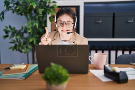 Téléchargez les photos : Woman with down syndrome working at the office wearing headset surprised with an idea or question pointing finger with happy face, number one - en image libre de droit