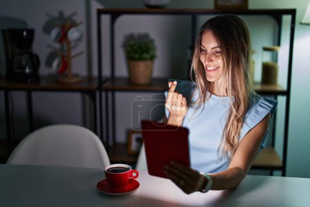 Téléchargez les photos : Young hispanic woman using touchpad sitting on the table at night doing money gesture with hands, asking for salary payment, millionaire business - en image libre de droit