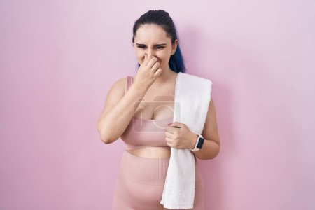 Foto de Young modern girl with blue hair wearing sportswear over pink background smelling something stinky and disgusting, intolerable smell, holding breath with fingers on nose. bad smell - Imagen libre de derechos