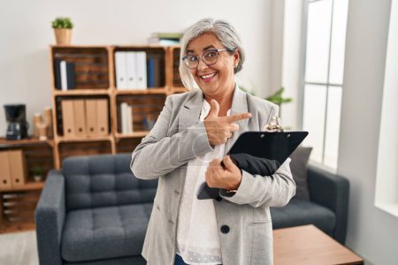 Foto de Mujer de mediana edad con el pelo gris en la oficina de consulta alegre con una sonrisa en la cara señalando con la mano y el dedo hacia el lado con expresión feliz y natural - Imagen libre de derechos