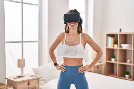 Téléchargez les photos : Jeune belle femme hispanique sportive utilisant des lunettes de réalité virtuelle debout à la chambre - en image libre de droit
