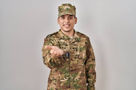 Foto de Joven árabe vestido con camuflaje uniforme del ejército sonriente alegre ofreciendo la palma de la mano dando asistencia y aceptación. - Imagen libre de derechos