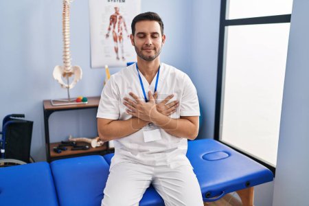 Foto de Young hispanic man with beard working at pain recovery clinic smiling with hands on chest with closed eyes and grateful gesture on face. health concept. - Imagen libre de derechos