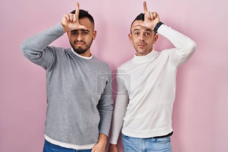 Téléchargez les photos : Couple homosexuel debout sur fond rose se moquant des gens avec les doigts sur le front faisant geste perdant moqueur et insultant. - en image libre de droit