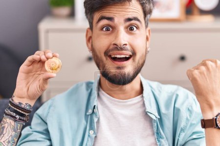 Foto de Young hispanic man with tattoos holding cardano cryptocurrency coin pointing thumb up to the side smiling happy with open mouth - Imagen libre de derechos