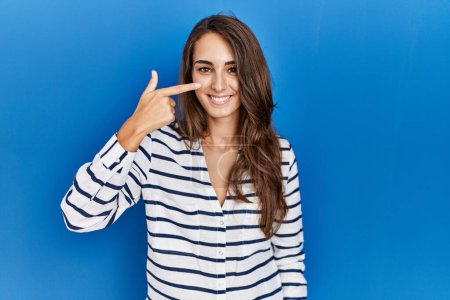Téléchargez les photos : Jeune femme hispanique debout sur fond bleu isolé pointant avec le doigt de la main vers le visage et le nez, souriant gai. concept de beauté - en image libre de droit
