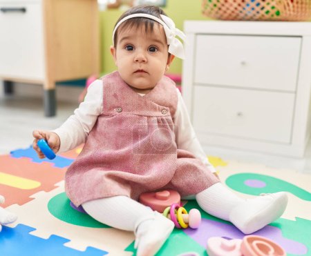 Téléchargez les photos : Adorable bébé hispanique assis sur le sol tenant jouet à la maternelle - en image libre de droit