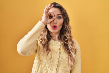 Photo for Young caucasian woman standing over yellow background doing ok gesture shocked with surprised face, eye looking through fingers. unbelieving expression. - Royalty Free Image