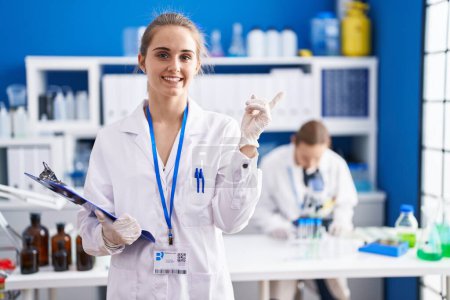 Foto de Blonde woman working at scientist laboratory smiling happy pointing with hand and finger to the side - Imagen libre de derechos